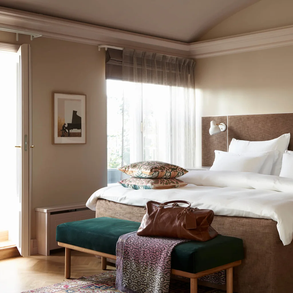 A neatly made bed with white bedding and decorative pillows sits in a cozy bedroom. A brown bag rests on a green bench at the foot of the bed.