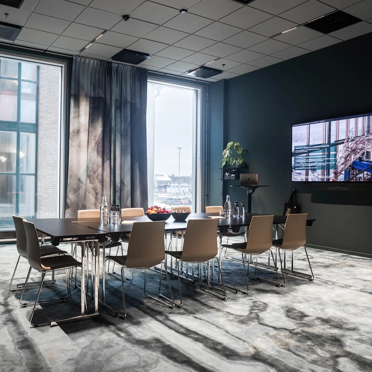 Ett konferensrum med ett rektangulärt bord omgivet av stolar, utrustat med vattenflaskor och fruktskålar. Ett stort fönster och TV-skärm som visar en interiör syns.