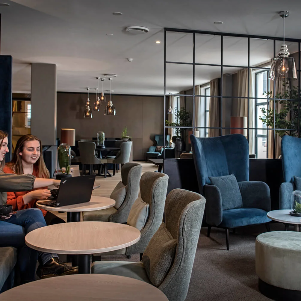Två kvinnor sitter och använder en bärbar dator i en stilfull hotelllobby med bekväma sittplatser och modern inredning.