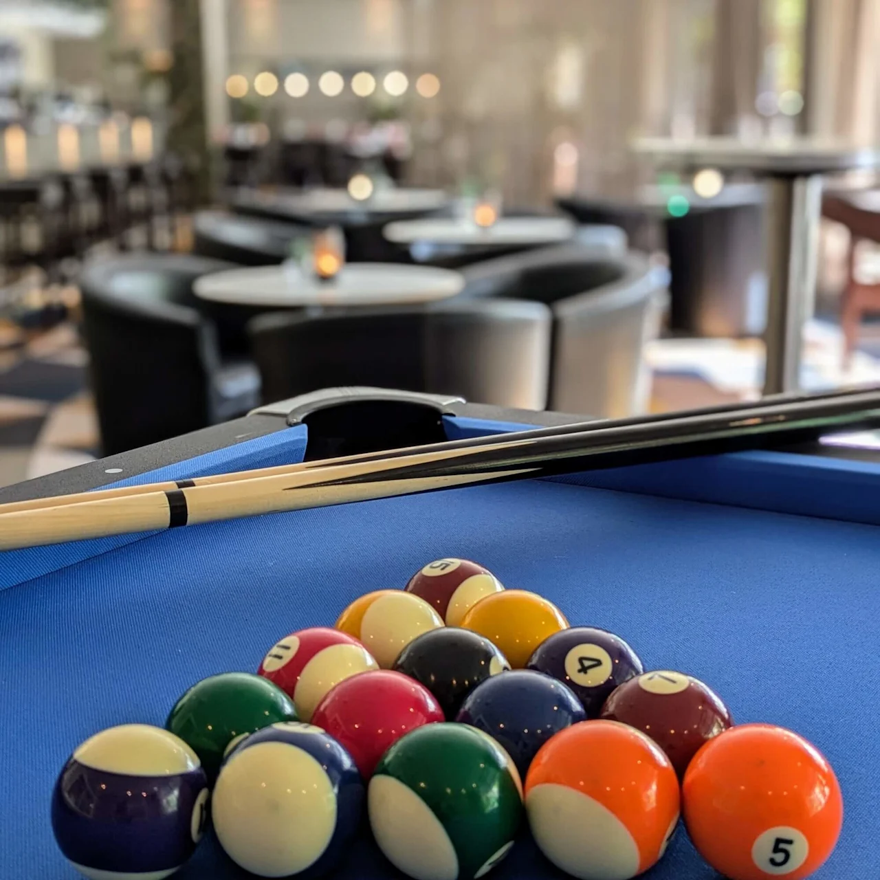 Biljardbollar arrangerade i en triangel på ett blått bord, köer vilandes ovanpå, i en modern lounge med runda bord och stämningsbelysning.