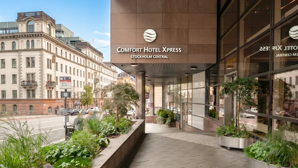The entrance of a modern hotel in a city with green plants and a classic building in the background.