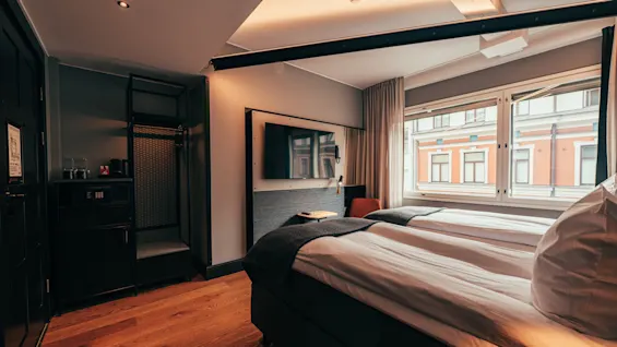 A cozy hotel room with two beds, a wall-mounted TV, and a large window offering a view of an urban building.