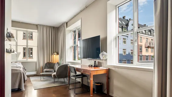 Et moderne hotelværelse har en seng, sofa og skrivebord med fjernsyn. Store vinduer tillader naturligt lys, og afslører en udsigt over farverige bygninger udenfor. Neutrale toner dominerer indretningen.