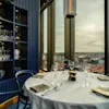 A restaurant table set for dining with a city view from large windows. Shelves with glassware are visible.