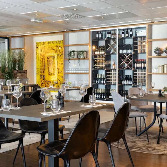 En restaurant med spiseplasser hvor bordene er elegant dekket med glass, omgitt av moderne stoler. En vinutstilling vises på bakveggen, sammen med dekorative planter og kunstverk.