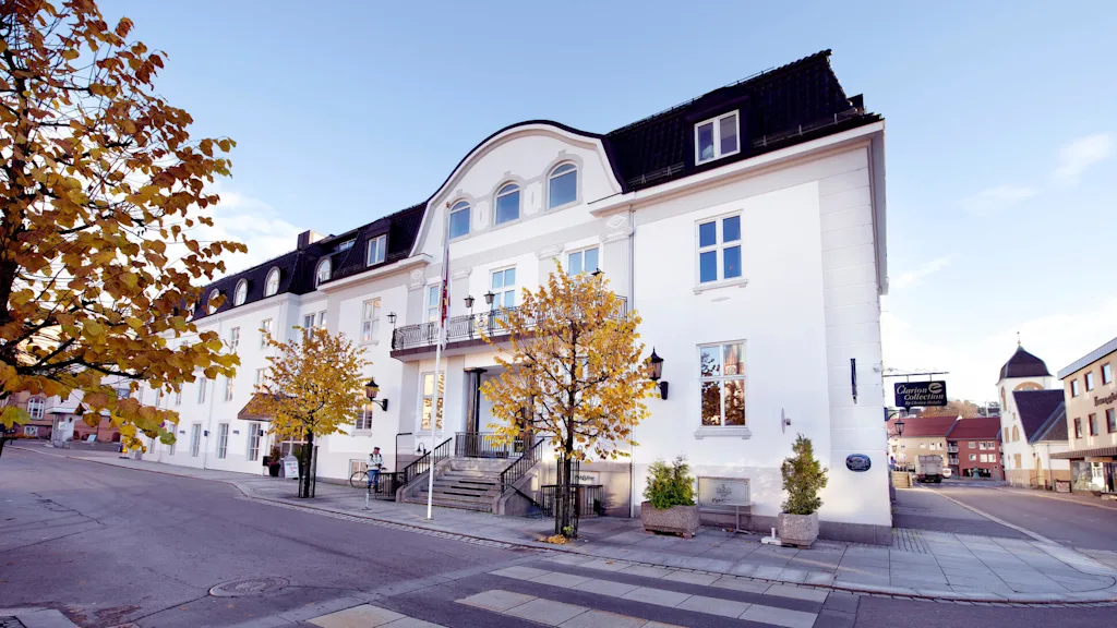 A white three-story building with black roof, balconies and autumn trees in a town setting during daytime.