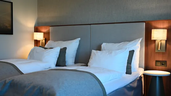 Et komfortabelt hotellrom med to pent oppredde senger, myk belysning og et nattbord, klart for gjester.