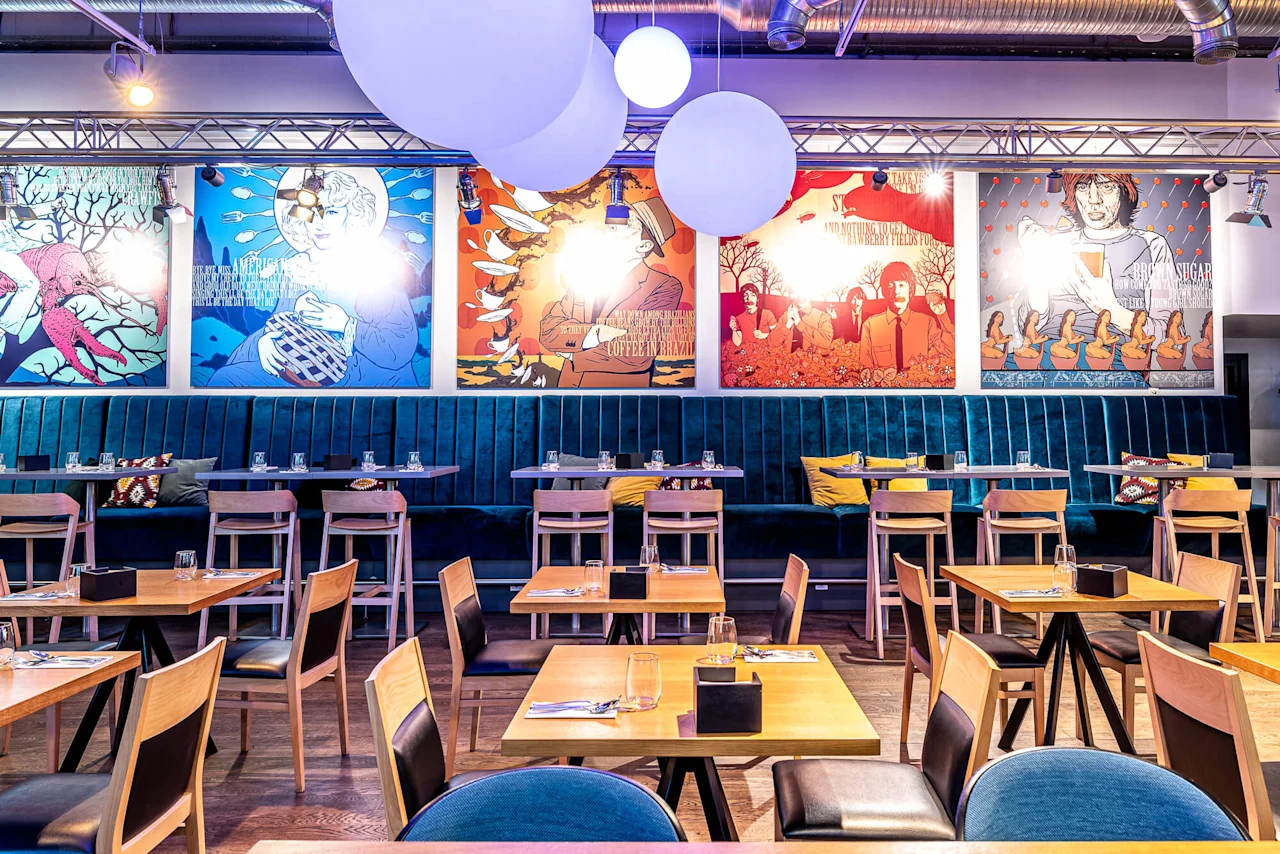 Restaurant interior featuring wooden tables and chairs, blue upholstered seating along the wall, and colorful artwork with abstract designs. Hanging globe lights add to the modern ambiance.