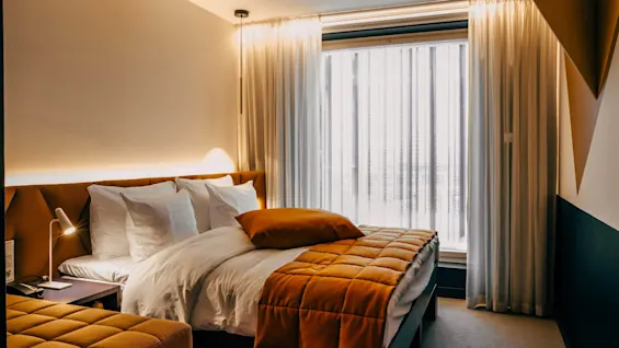 A neatly made bed with white pillows and an orange quilt is in a modern hotel room with warm lighting, beside a window with sheer curtains, creating a cozy atmosphere.