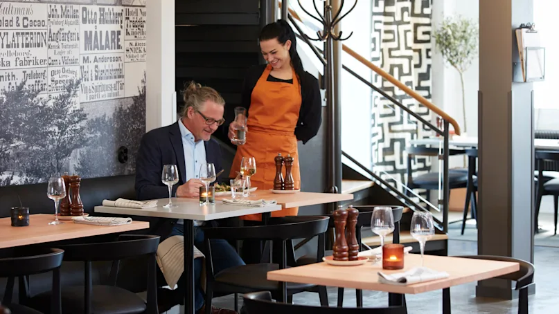 A man sits enjoying a meal as a waitress stands by, both smiling in a restaurant with modern decor.