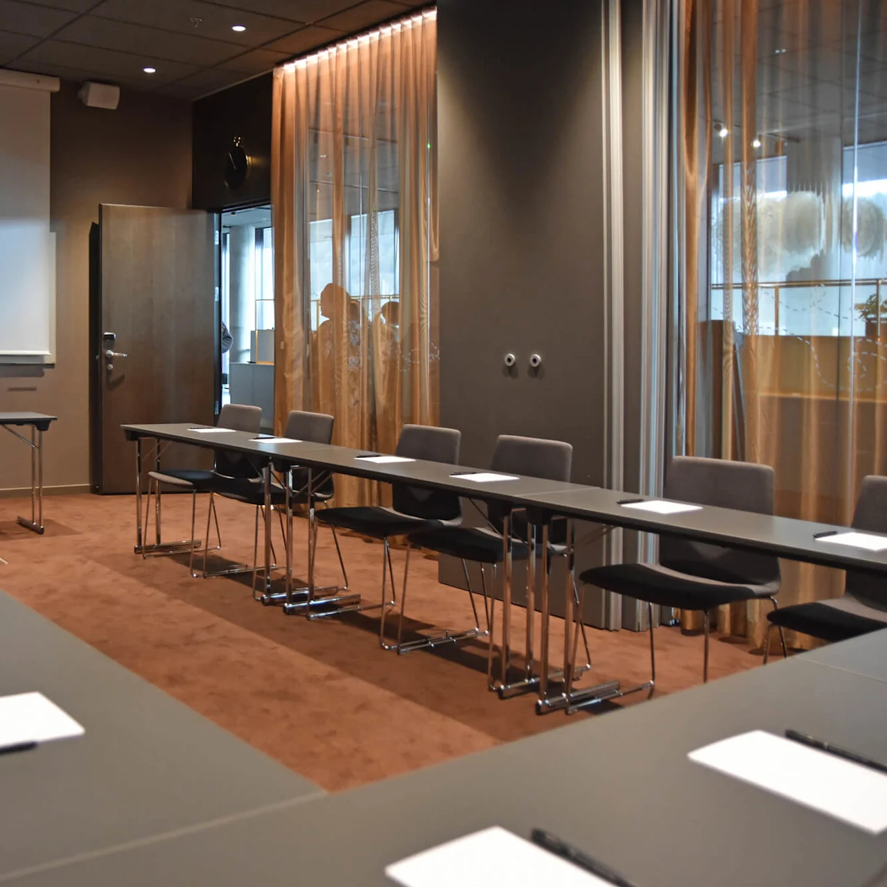 Konferensrum med U-format bord och stolar; anteckningsblock på bordet, projektorskärm på väggen, gardiner vid fönstren skapar en affärsmötesmiljö.