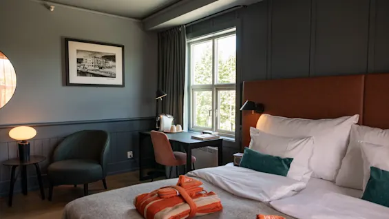 Et pænt arrangeret hotelværelse har en seng med hvidt sengetøj og orange badekåber, ved siden af et vindue. Et skrivebord med lampe og stol giver arbejdsplads ved siden af sengen.