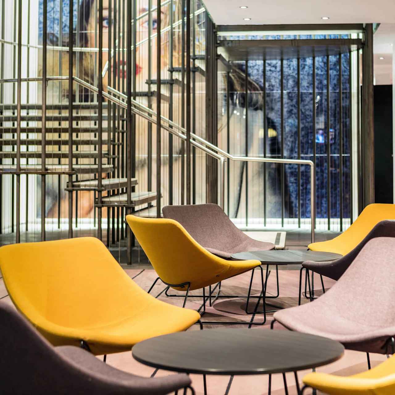 Yellow and brown lounge chairs surround small tables, set in a modern, open space with metal railings and a staircase, under soft lighting.