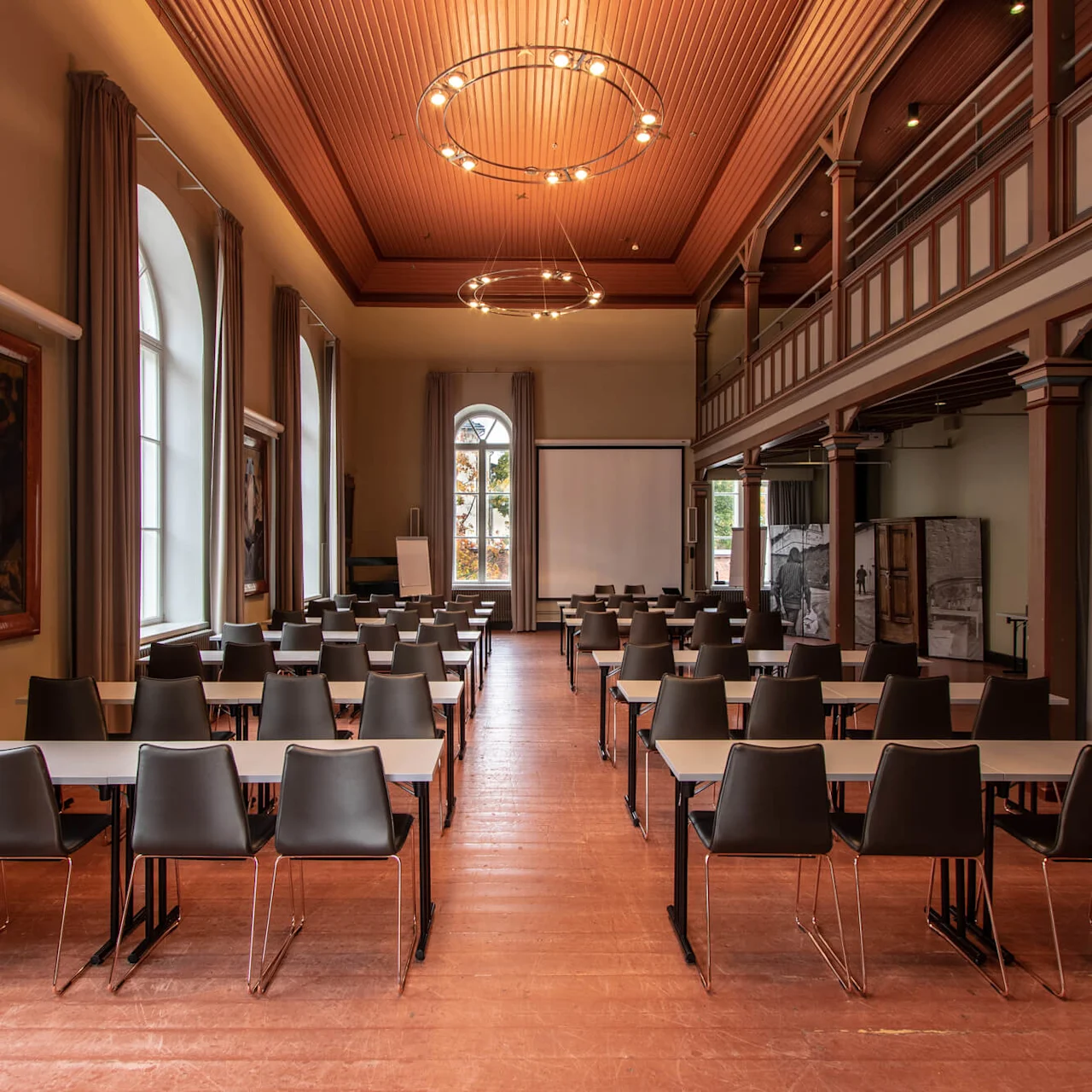 Rows of black chairs facing a whiteboard in a spacious room with wood-paneled walls, large windows, and paintings.