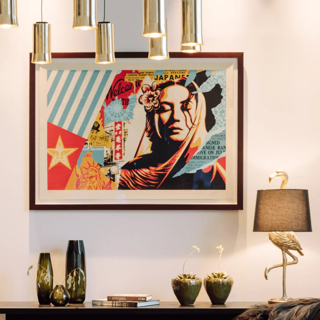 A framed colorful artwork hangs on the wall, depicting a woman with floral elements and text like "Welcome to" and "JAPANESE BAN." Below, decorative vases and a lamp sit on a table.