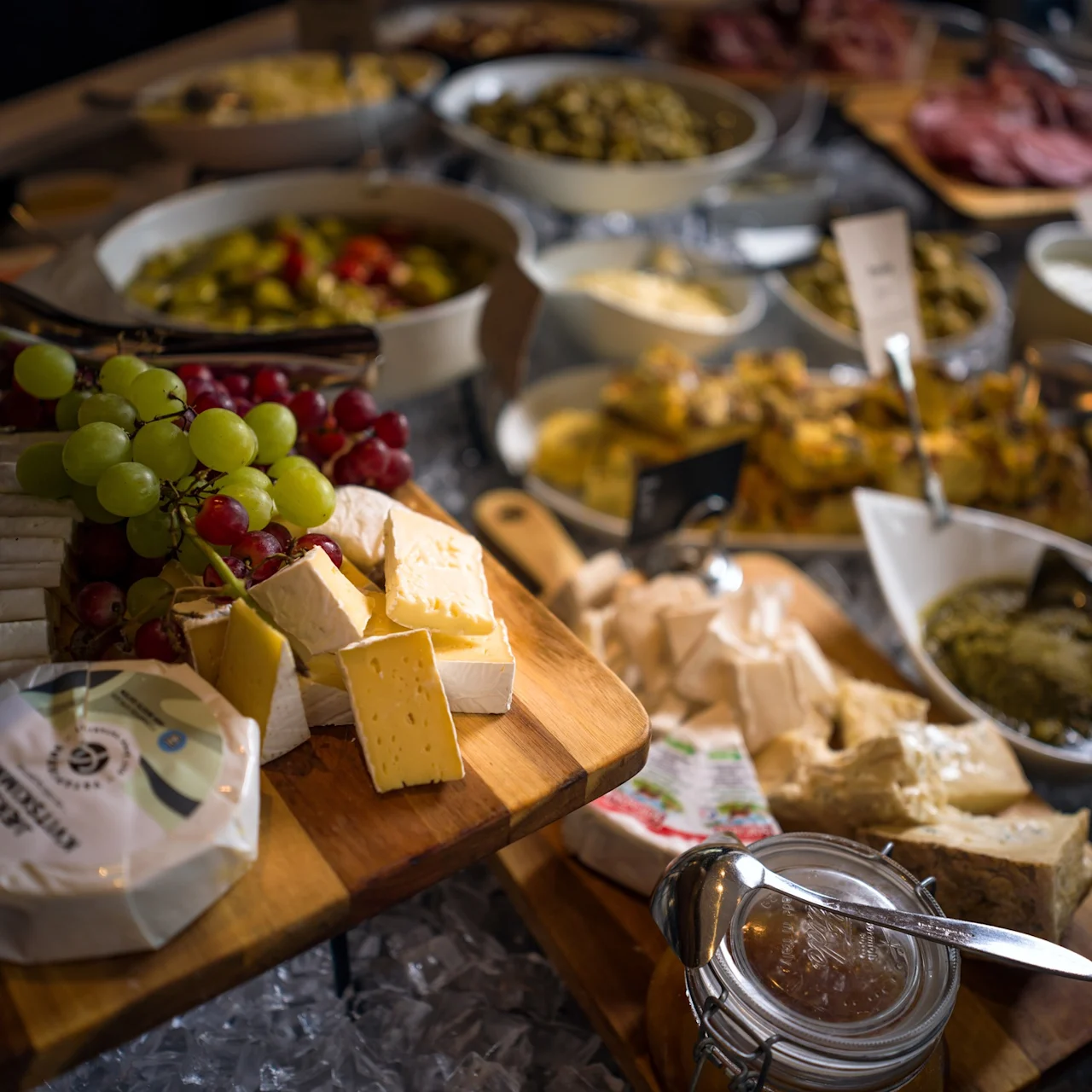 En ostbricka upptar förgrunden, med olika ostar, röda och gröna vindruvor, samt en burk sylt. I bakgrunden finns olika rätter utspridda över ett buffébord.