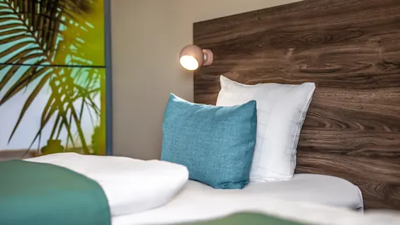 A bed with white and teal pillows and blankets is placed against a wooden headboard. A wall-mounted light is above the pillows and a leafy plant print decorates the room.