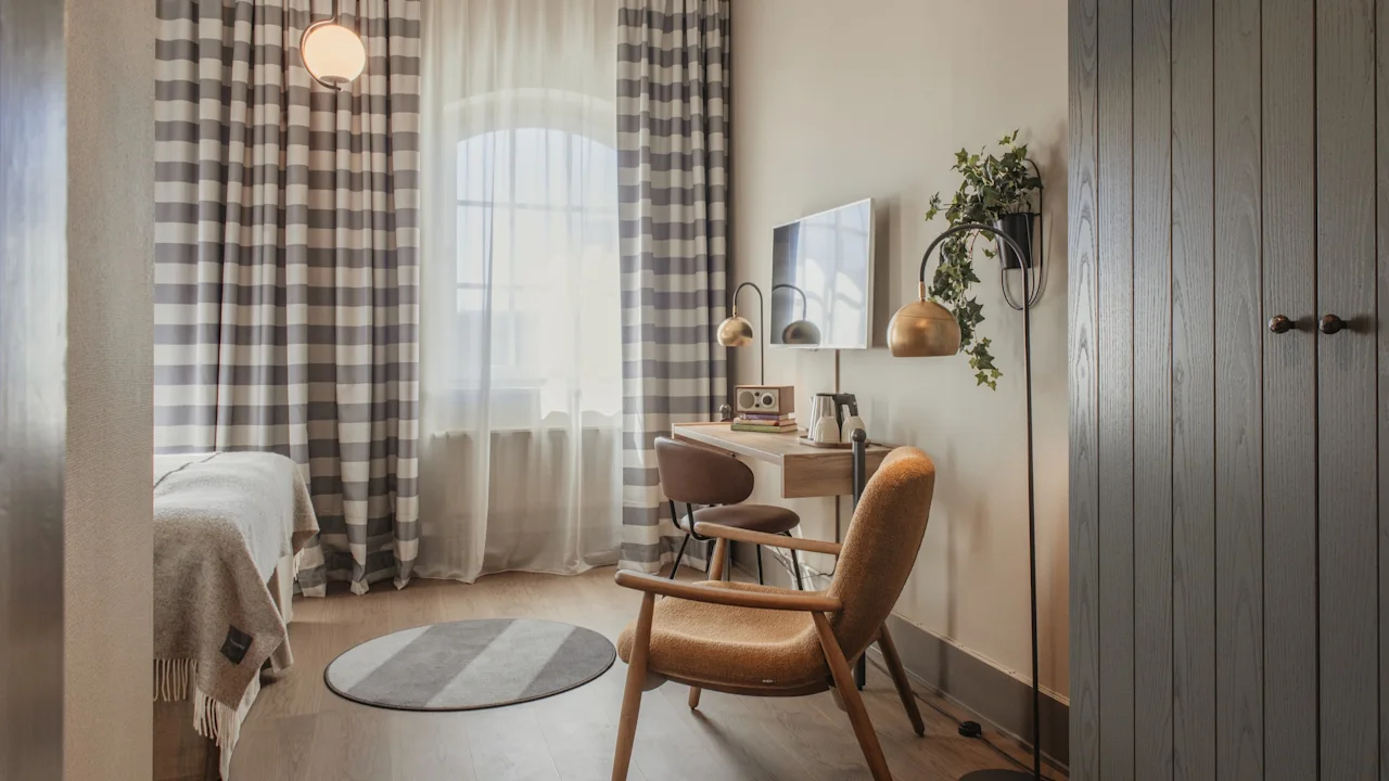 Ett mysigt hotellrum med en säng, en skrivbordsplats och en bekväm fåtölj, med rutiga gardiner och varm belysning.