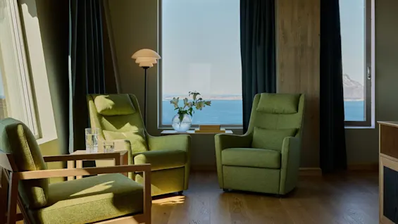Green armchairs face each other beside a wooden table with glasses, a vase of flowers, and a lamp. They overlook a large window showing a sea view.