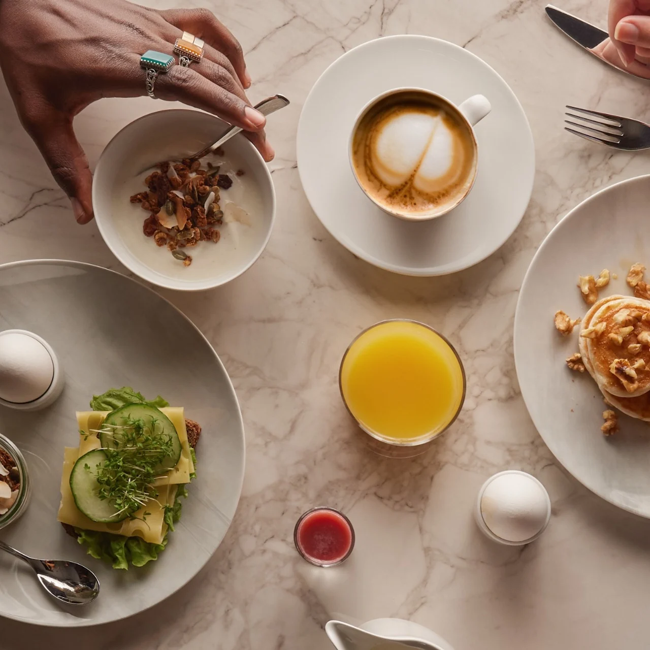 Ett varierat frukostbord: en person rör om yoghurt med granola, en annan håller bestick. Tallrikar med ägg, grönt, rostat bröd, grapefrukt, en croissant; cappuccino, juice och sylt på marmorbord.