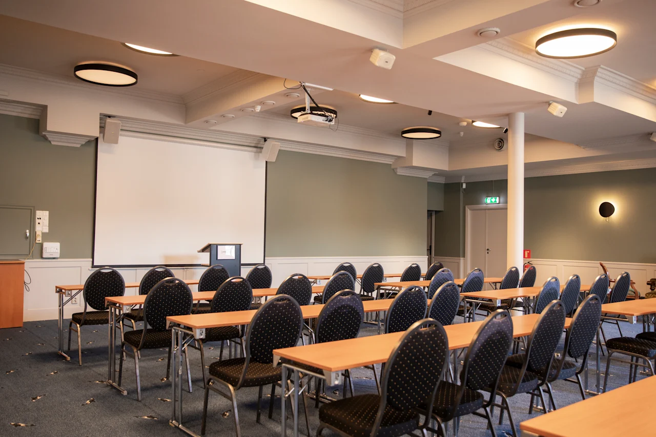 Et konferencelokale med rækker af borde og stole vendt mod en stor projektorskærm og et podium, klar til en præsentation.