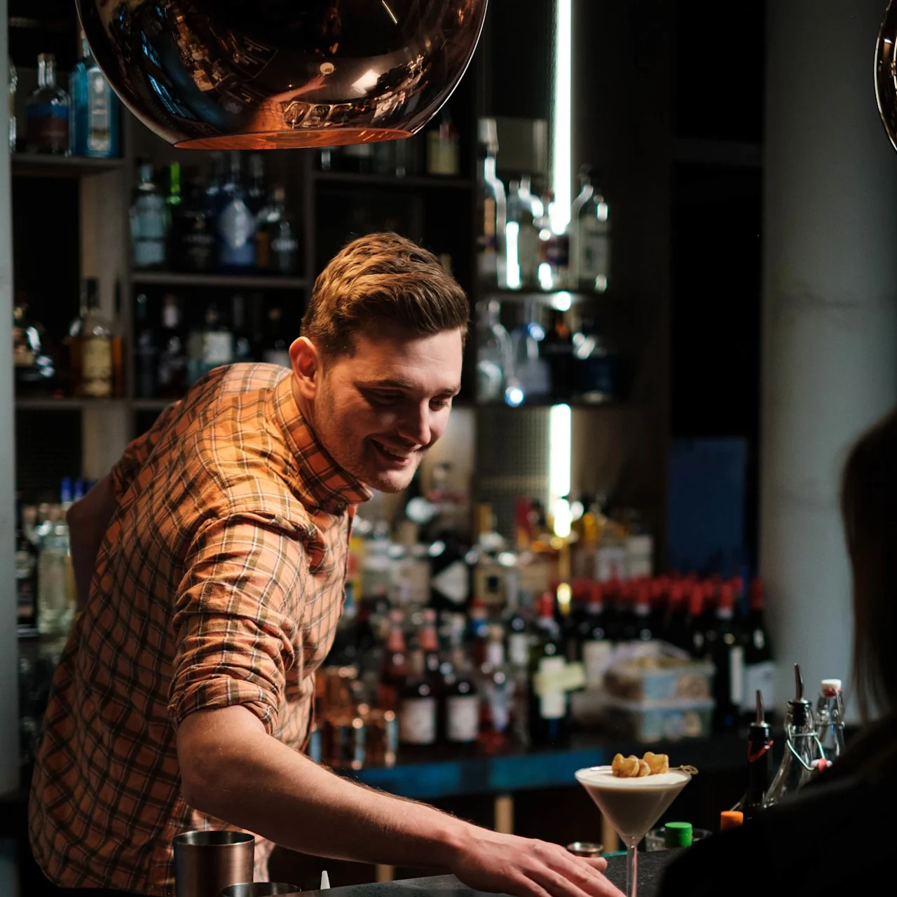 A smiling bartender in a plaid shirt serves a cocktail at a dimly lit bar, with shelves full of various liquor bottles in the background.