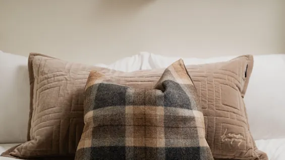 En hyggelig seng med ternede og teksturerede puder, oplyst af en varm væglampe, inviterer til afslapning.