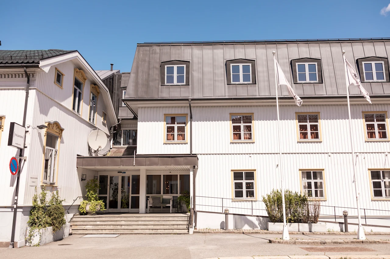 En trevånings vit byggnad står med metalltak, omgiven av tre flaggstänger och grönska. Ingången har en liten trappa, under klarblå himmel.