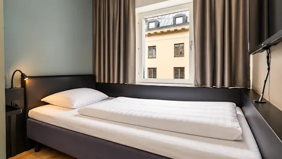 A single hotel room featuring a single bed placed under a window with a view of a building.