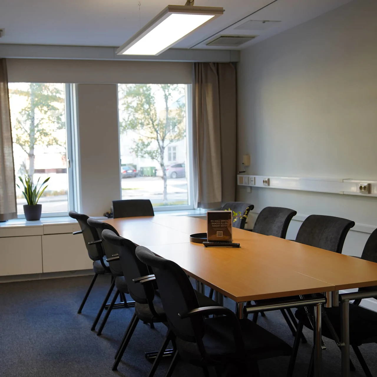 Ett konferensrum med ett långt träbord omgivet av svarta stolar. En whiteboard hänger på väggen, och stora fönster med gardiner släpper in dagsljus. En skylt lyder "FÅ KLARA SVAR - ROBERT BULLER".