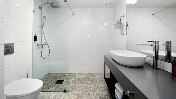 A modern bathroom features a glass-enclosed shower with a rainfall showerhead, wall-mounted toilet, large mirror, and sleek sink on a concrete countertop, surrounded by white tiles and grey stone floor.
