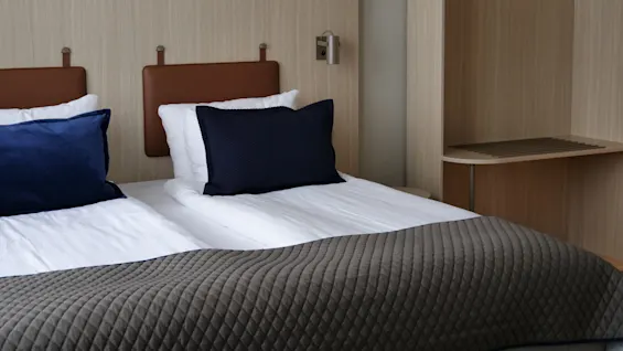 A neatly made bed with blue and white pillows rests against a light wooden headboard. Nearby, a wooden luggage rack enhances the modern, minimalist room decor.