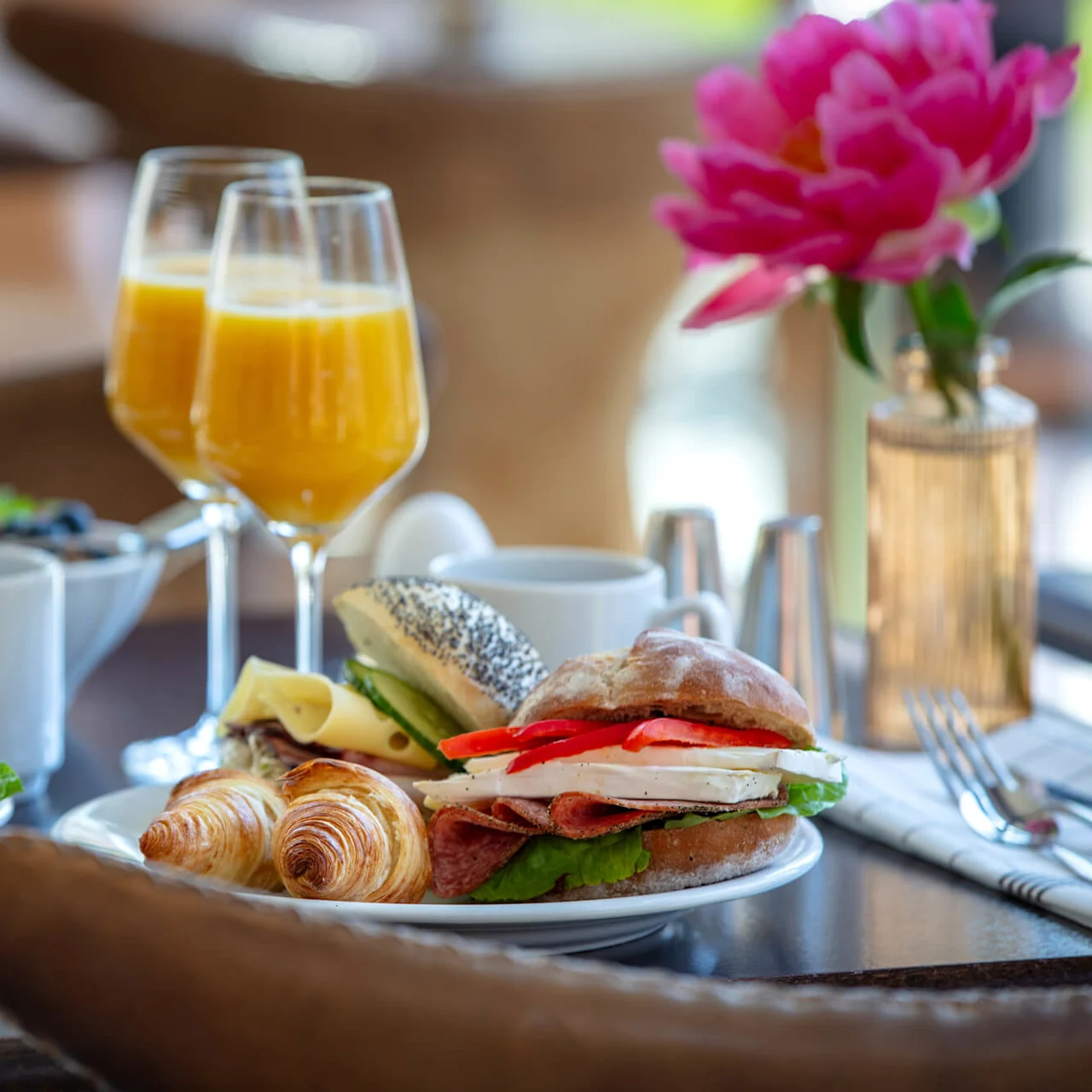 A breakfast spread with orange juice, coffee, pastries, and a sandwich, arranged on a table with a pink flower in the background.