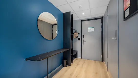 En moderne hotellromsinngang med en blå vegg, et rundt speil, et elegant konsollbord og et lyst tregulv som fører til en lukket dør.