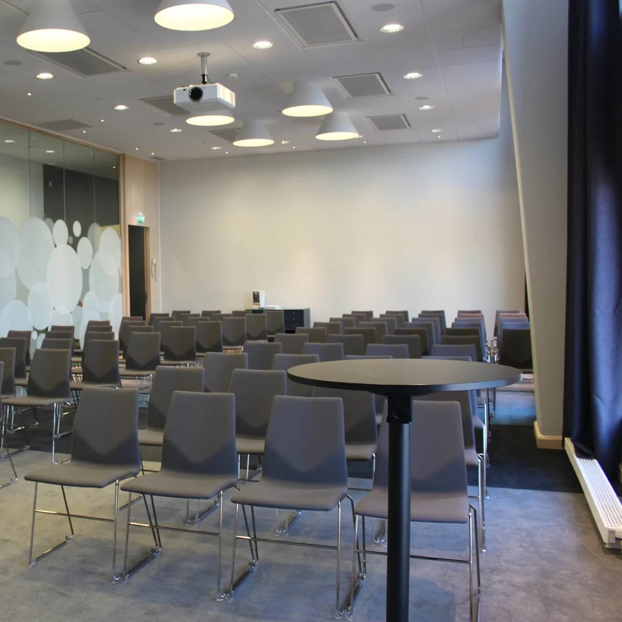 Rækker af tomme grå stole vender mod en vægprojektor i et moderne konferencelokale. Frostet glasvægge med cirkulære mønstre flankerer rummet, oplyst af loftslamper.