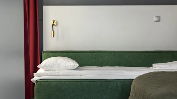 Ett mysigt, modernt sovrum med en prydligt bäddad säng i grönt mot en enkel sänggavel och en vägglampa i guld. Röda gardiner ger värme, medan en bunt handdukar antyder en välkomnande atmosfär.