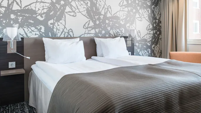 En pent dobbeltseng med hvite puter og et grått teppe hviler mot en vegg med abstrakt mønster. En oransje stol og et vindu med beige gardiner flankerer sengen.