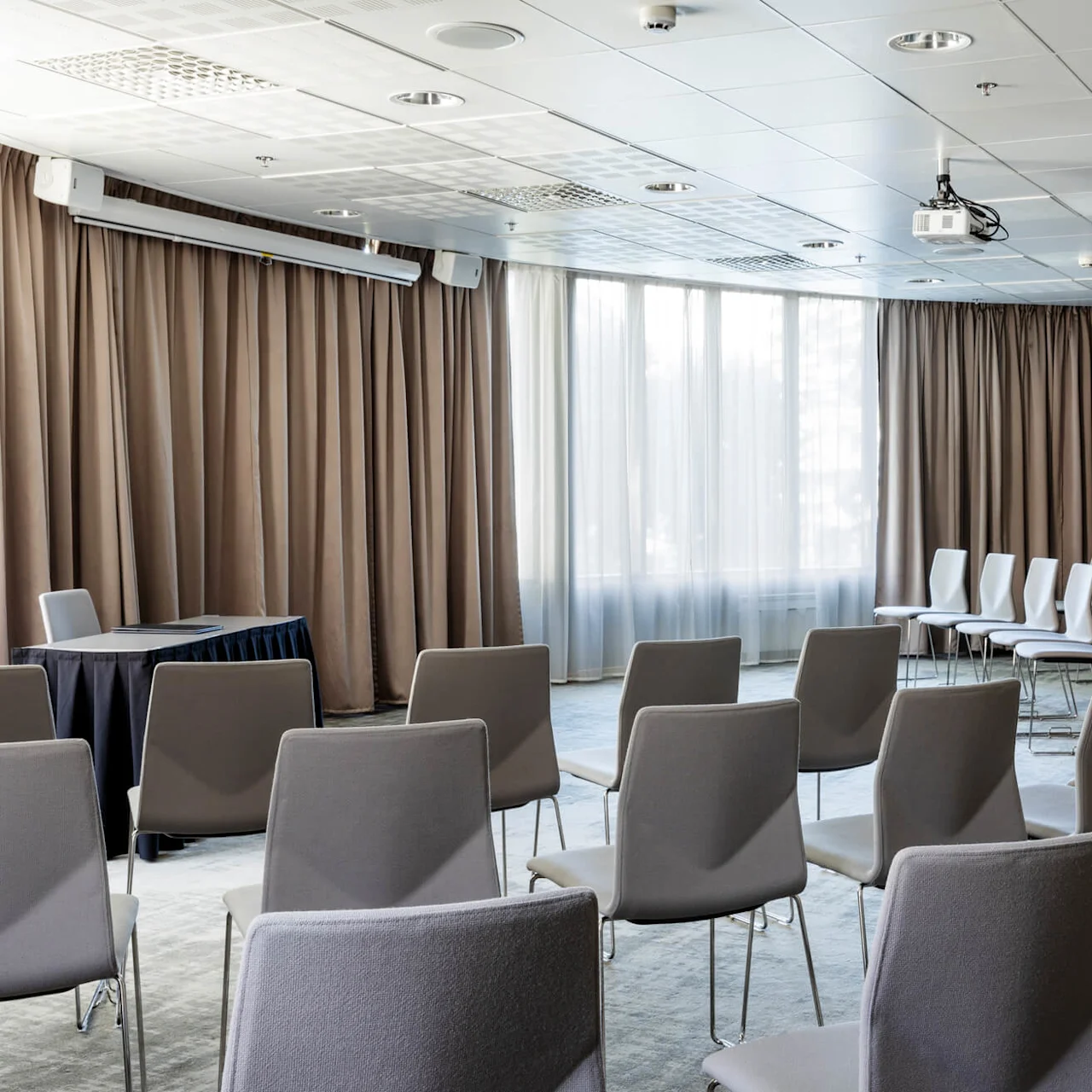 Rows of gray chairs are arranged in a semi-circle, facing a table with a dark cloth. A projector hangs from the ceiling in a room with large windows and brown curtains.