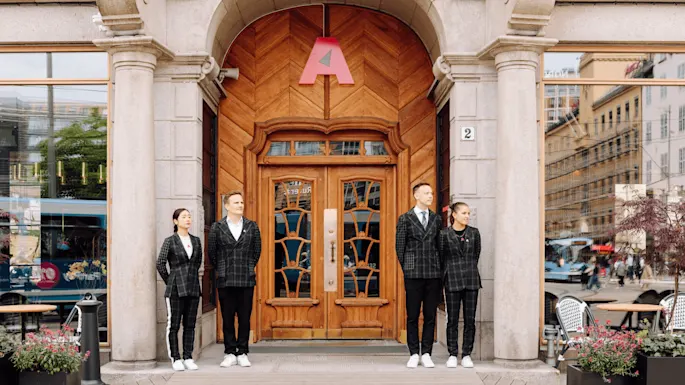 Four people stand outside a large wooden door with an arch, wearing matching plaid suits. There is a large red letter "A" above the door. Nearby, glass windows reflect a busy street. A plaque shows the number "2."