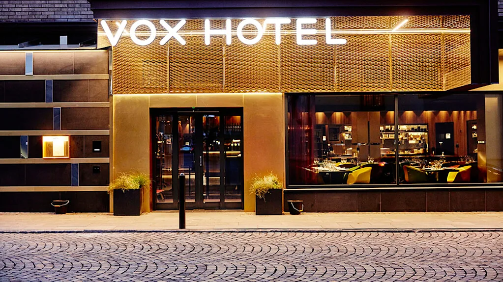VOX HOTEL-skiltet lyser klart på en bygningens facade. Nedenfor fører glasdøre ind i et varmt oplyst interiør med spiseborde. Brostensbelagt gade foran.