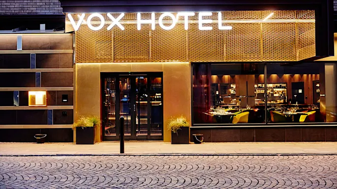 VOX HOTEL-skilt lyser sterkt på en husfasade. Under leder glassdører inn i et varmt opplyst interiør med spisebord. Brosteinsgate i front.