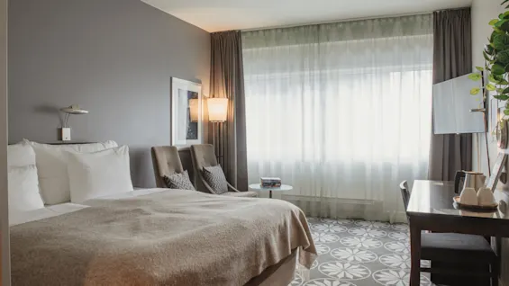 Ett bekvämt och modernt hotellrum med en stor säng, sittgrupp och ett skrivbord, badat i naturligt ljus från ett stort fönster.