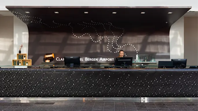 A long reception desk with computers and a staff member stands in a modern lobby. Behind, a black wall features dotted artwork and the text "CLARION HOTEL BERGEN AIRPORT."
