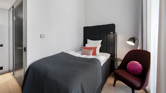 A neatly made bed with black headboard and white pillows rests against a gray wall. Beside it, a red chair with a purple cushion sits under a lit floor lamp.