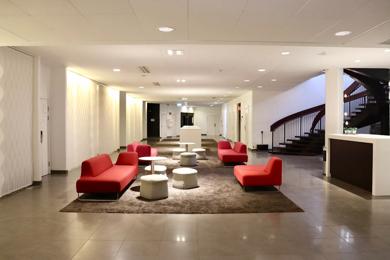 Red sofas sit on a brown rug, surrounded by white stools in a modern lobby with a spiral staircase and overhead lighting.