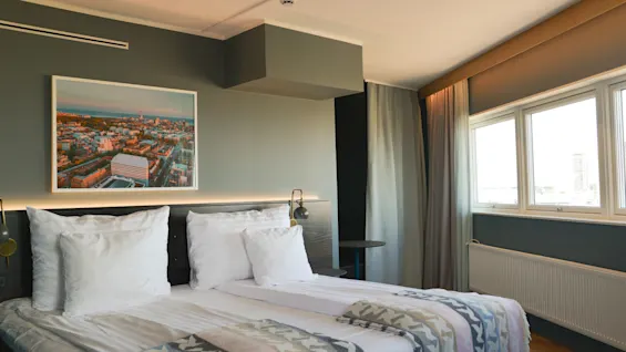 En pænt redt seng med hvide puder og mønstrede tæpper står i et hotelværelse. Et bylandskabsfoto hænger over, med store vinduer og gardiner til højre.