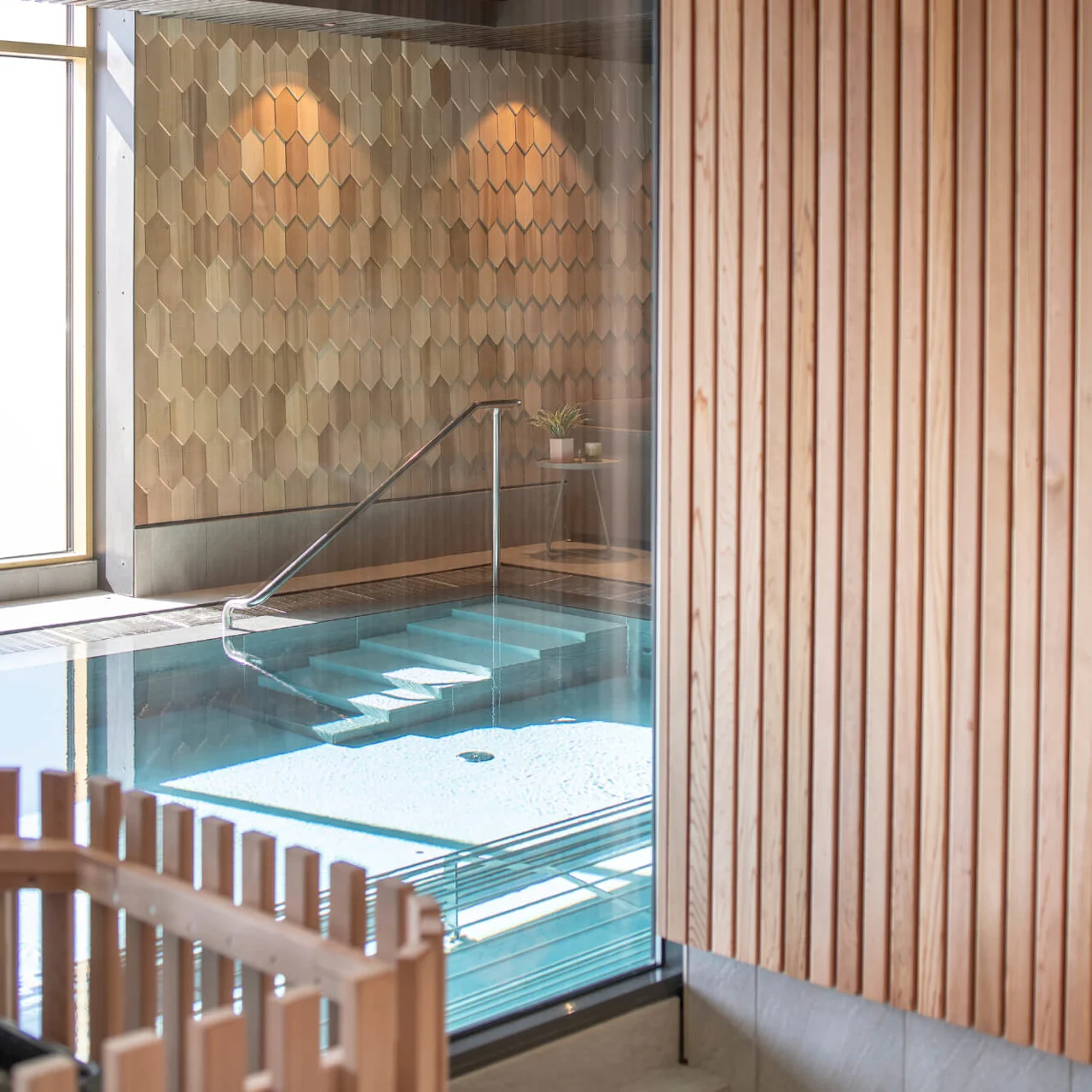 A small indoor swimming pool with steps leading in, surrounded by wooden paneling and honeycomb-patterned tiles, situated in a modern, sunlit room.