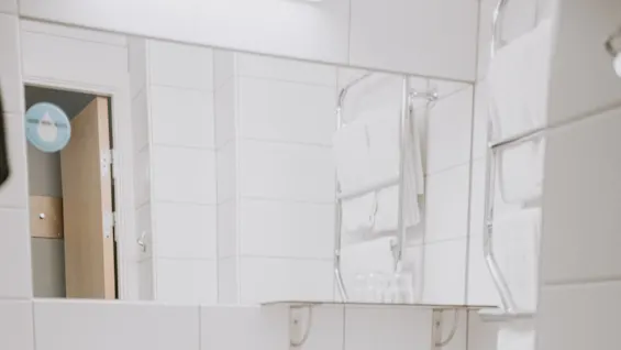 Håndvask med sæbedispenser står mod en hvid flisebelagt badeværelsesvæg; et spejl reflekterer håndklæder på en holder. Der er et toilet i nærheden og en metalaffaldsspand nedenfor. 