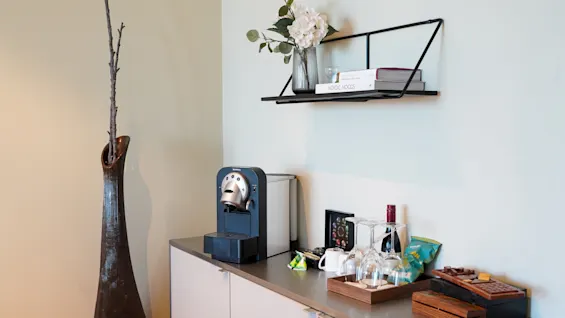 A coffee machine sits on a countertop beside wine glasses and snacks, with a tall vase holding branches nearby. A wall shelf holds books and flowers. "NORDIC MOODS" is visible.