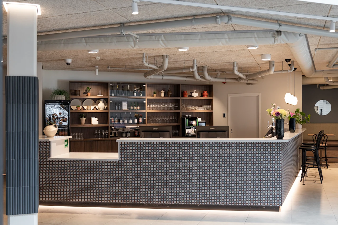A modern hotel reception or bar area with a patterned counter, shelves filled with bottles and glasses, and floral decorations.